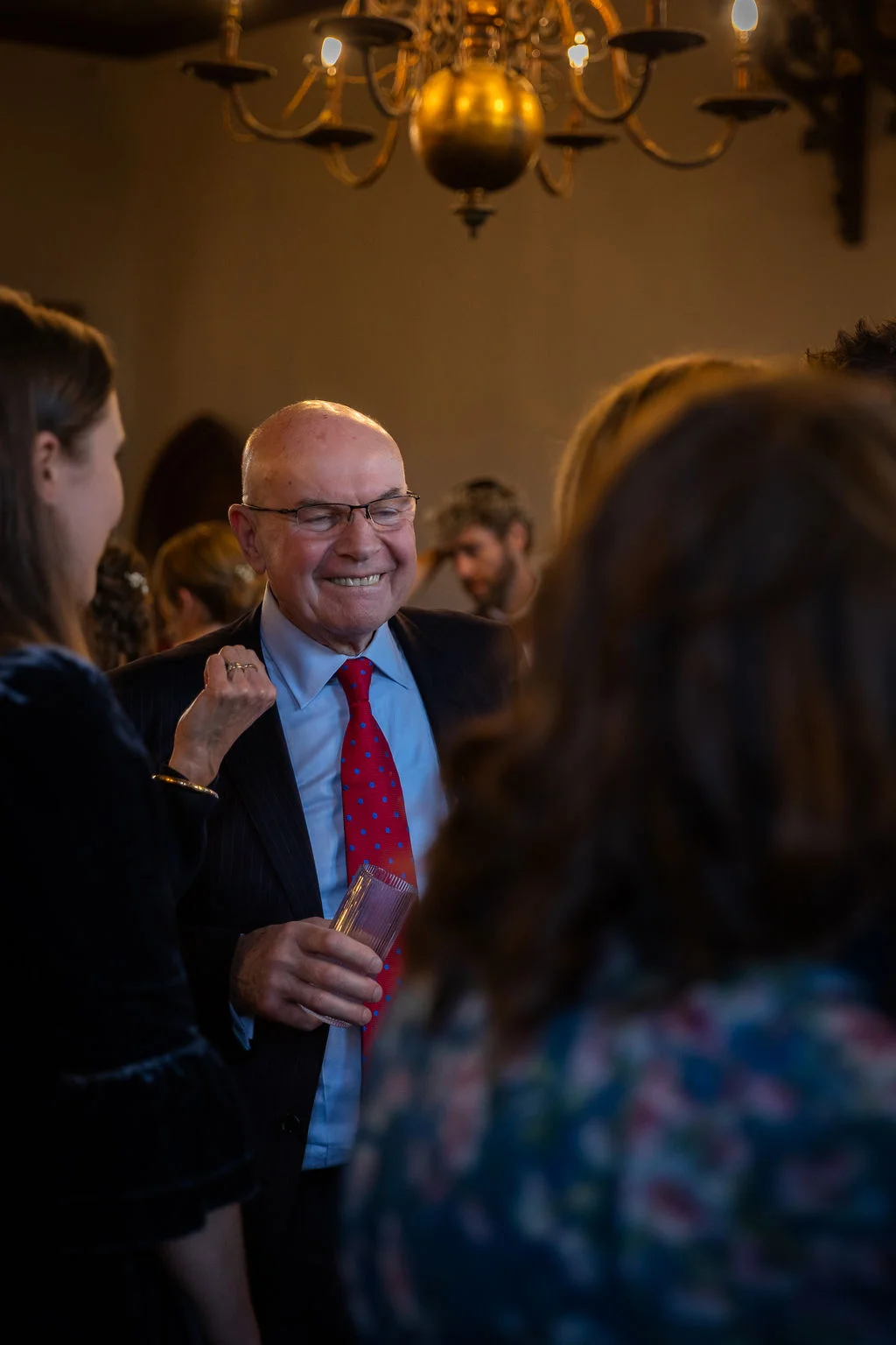 A smiling older male wedding guest wearing glasses, a dark suit, and a red patterned tie is captured chatting with guests during a reception under a warm chandelier.