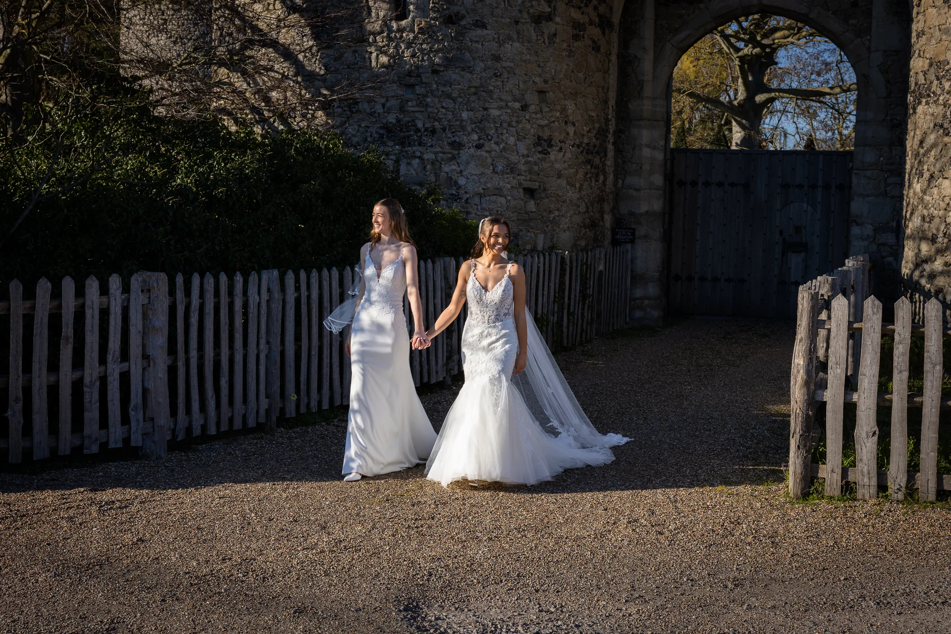 Two smiling brides holding hands as they walk together outdoors in front of an ancient stone castle archway on a sunny day.