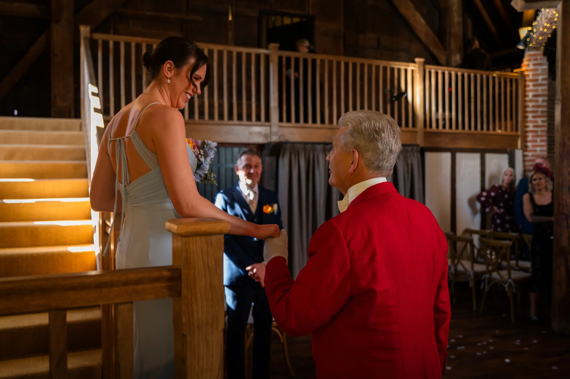 A smiling bride in a dusty blue dress holds hands with her father in a red jacket inside a rustic barn venue.