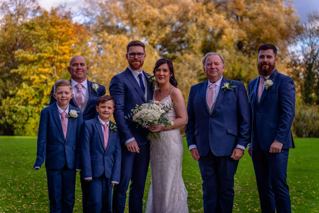 A bridal party group portrait poses outdoors on a green lawn against a backdrop of autumn trees with golden foliage.