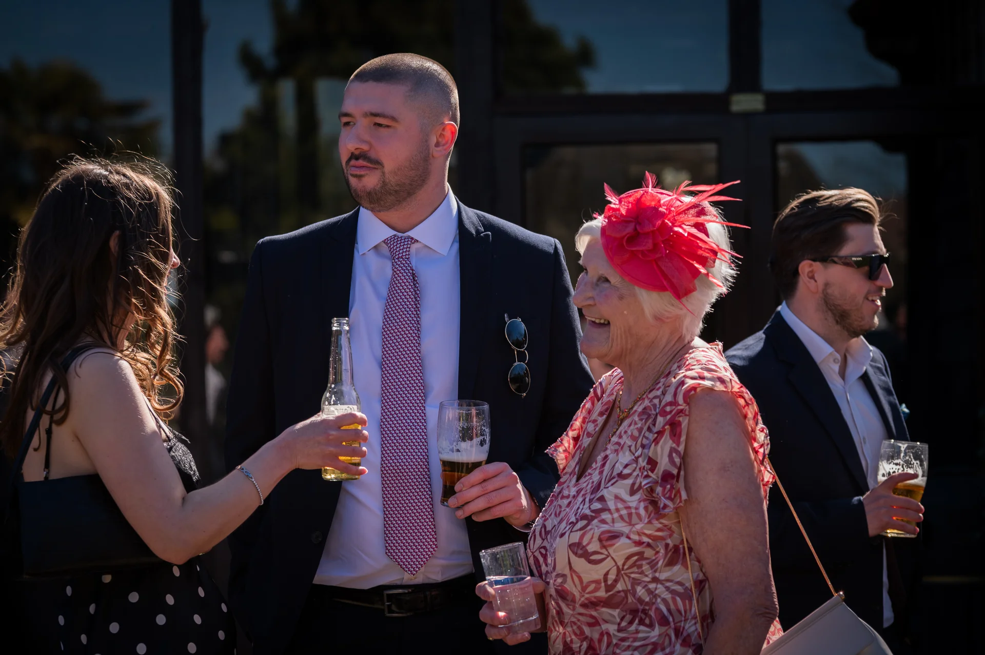 Wedding guests mingle and chat happily outdoors during a sunny drinks reception, holding drinks and wearing formal attire and a bright fascinator.