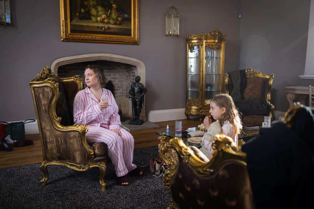 A woman wearing matching pink striped pyjamas sits in a gilded armchair holding a glass while a child sits nearby in an elegant sitting room.