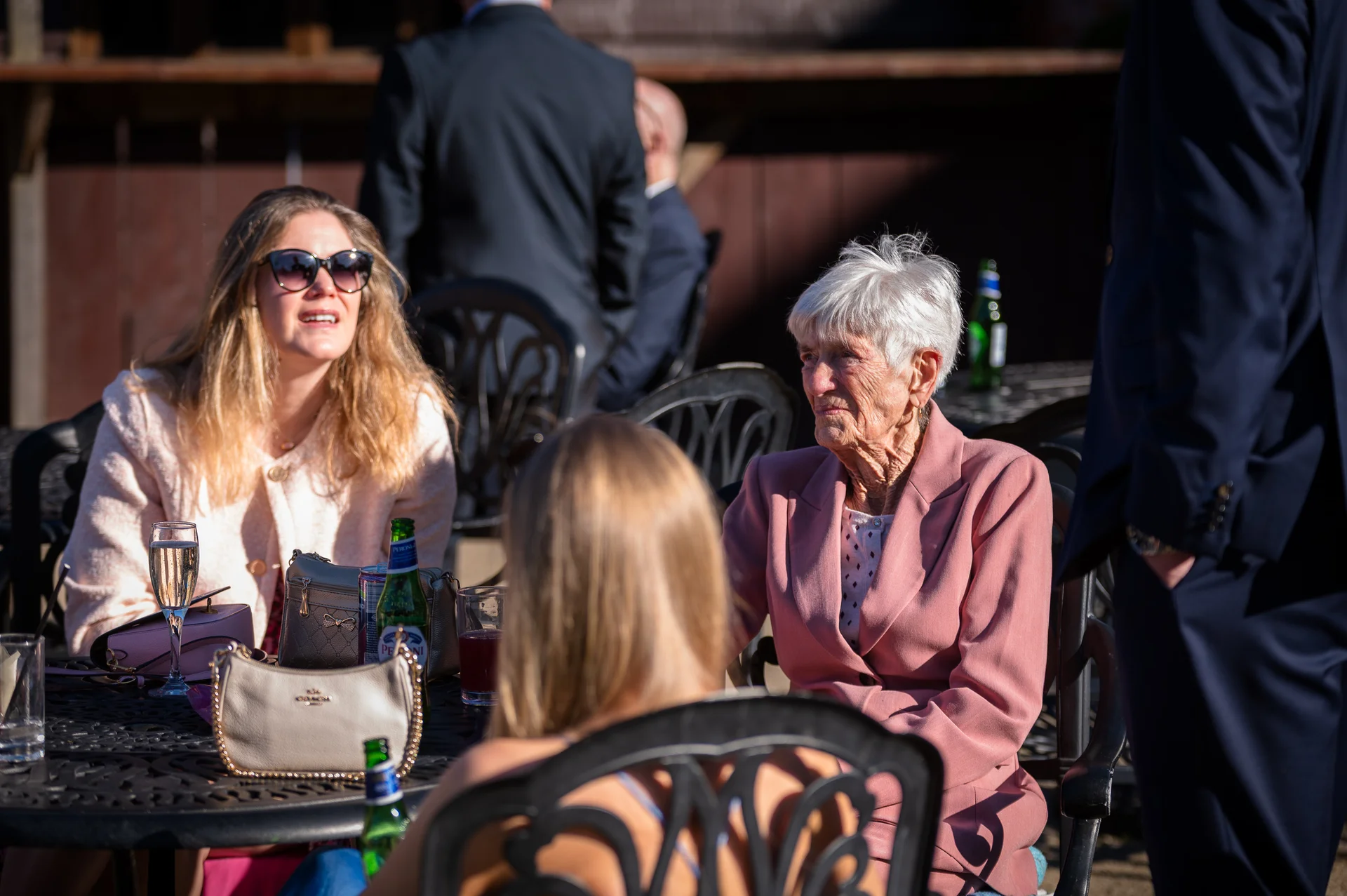 An elderly woman in a pink blazer and wedding guests enjoy a sunny outdoor drinks reception, seated at a black metal table.