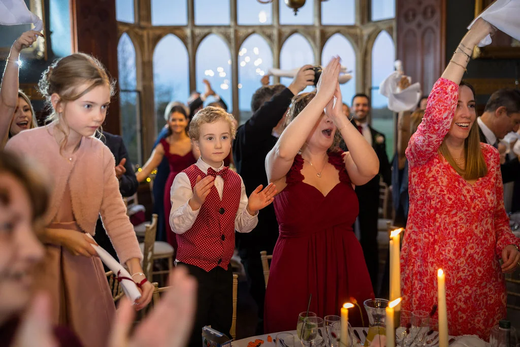 Wedding guests enthusiastically cheer and wave napkins during the wedding speeches inside a grand country house dining room with large arched windows.