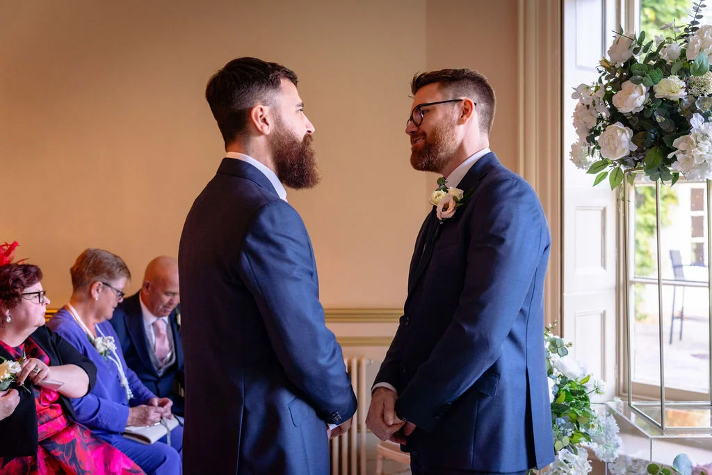 Two grooms in navy suits share a tender moment looking into each other's eyes during their indoor wedding ceremony.