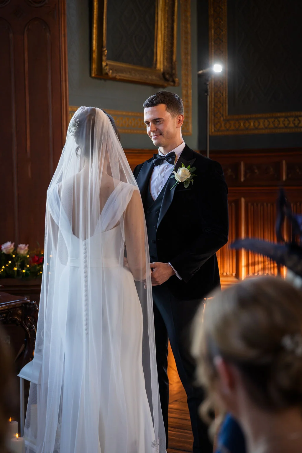 A groom looks adoringly at his bride standing opposite him at the altar inside an elegant wood-panelled wedding venue.