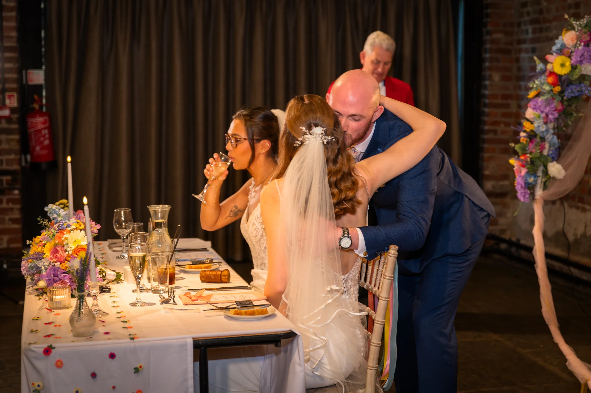 A groom leans over the top table to kiss his bride during the wedding breakfast while a guest drinks champagne nearby.
