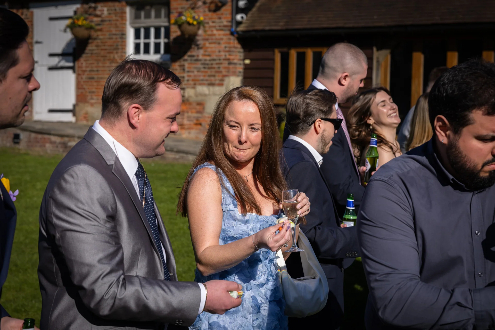 Wedding guests mingle outdoors in the sunshine during a drinks reception, holding glasses of champagne and enjoying the celebration.