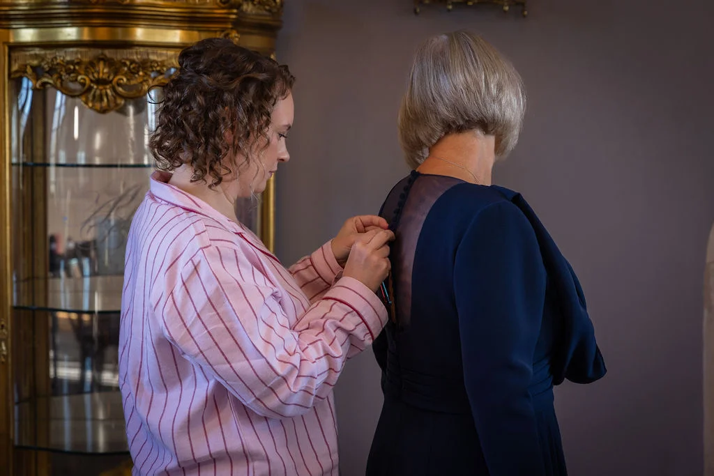 A woman wearing pink striped pyjamas carefully fastens the buttons on the back of a navy dress for another woman during morning wedding preparations.