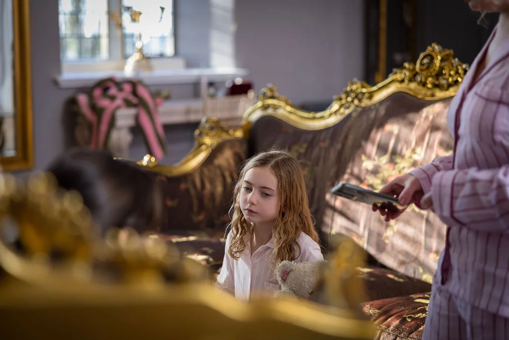 A young flower girl wearing matching pyjamas sits on an ornate gold and brown sofa inside a stately home during wedding morning preparations.