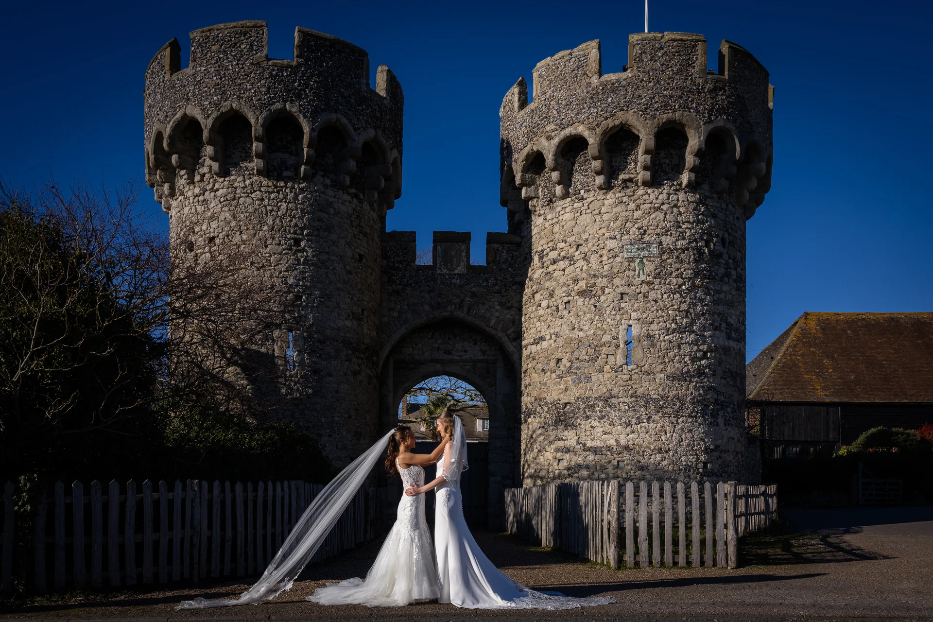 Two brides in wedding dresses stand embracing outside a historic stone castle gate under a bright blue sky.