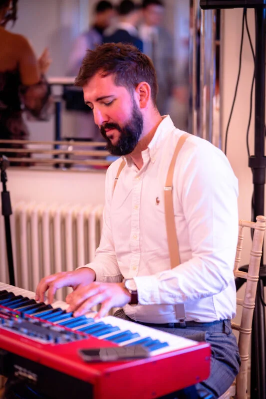 A bearded musician wearing a white shirt and braces plays a keyboard during a wedding celebration.
