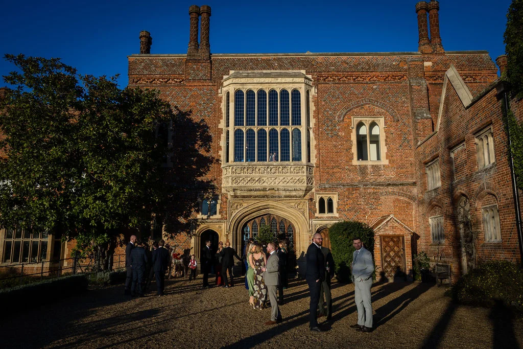 Wedding guests mingle in the sunlit cobbled courtyard of a grand historic red-brick country house during the drinks reception.