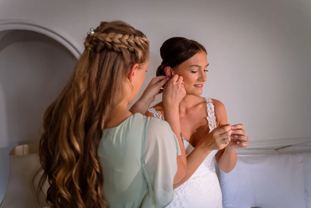 A bridesmaid helps the bride put on her earrings during wedding morning preparations in a bright room.
