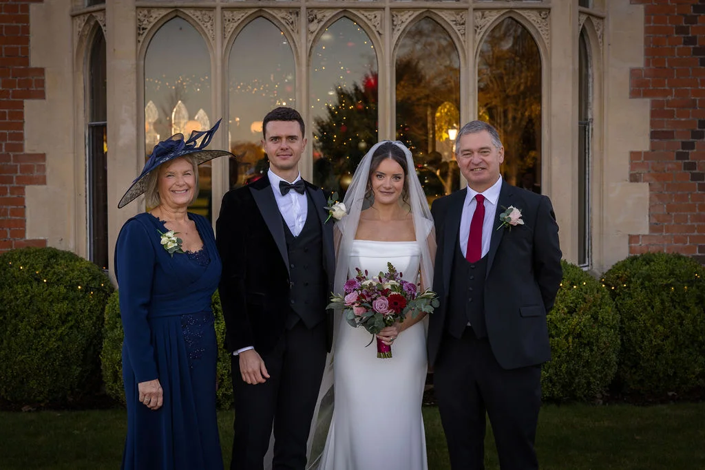 A newlywed couple stands outside a grand country house with family members, posing before arched windows with topiary bushes in the foreground.