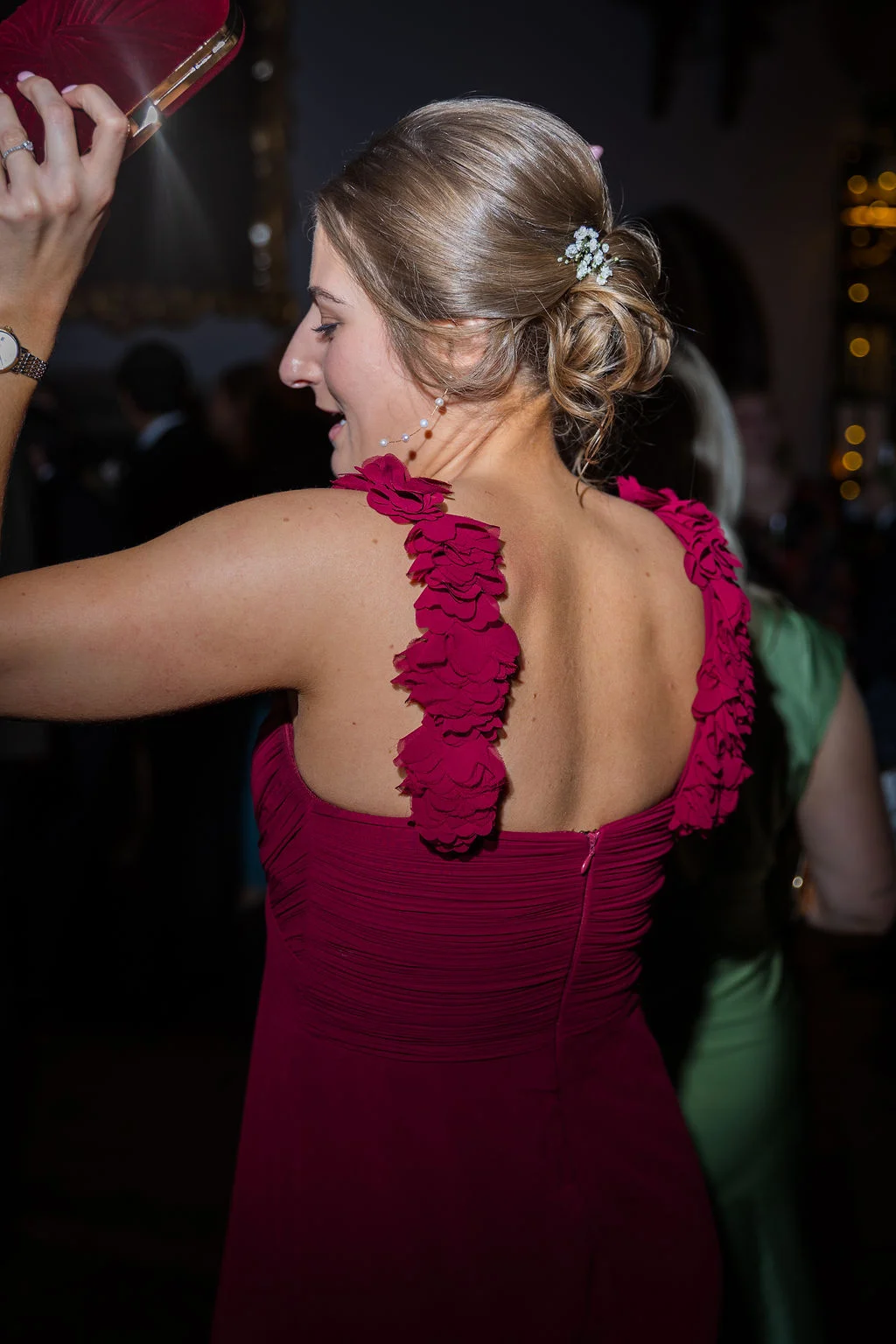 A wedding guest wearing a ruched burgundy dress with floral strap details and holding a clutch dances at the evening reception.