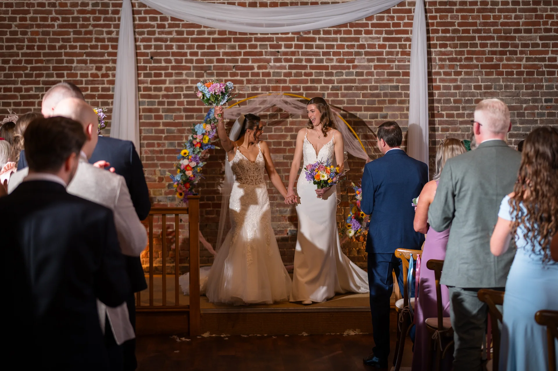 Two smiling brides joyfully hold hands and raise their bouquets as they walk back down the aisle after their indoor ceremony in a rustic brick barn.
