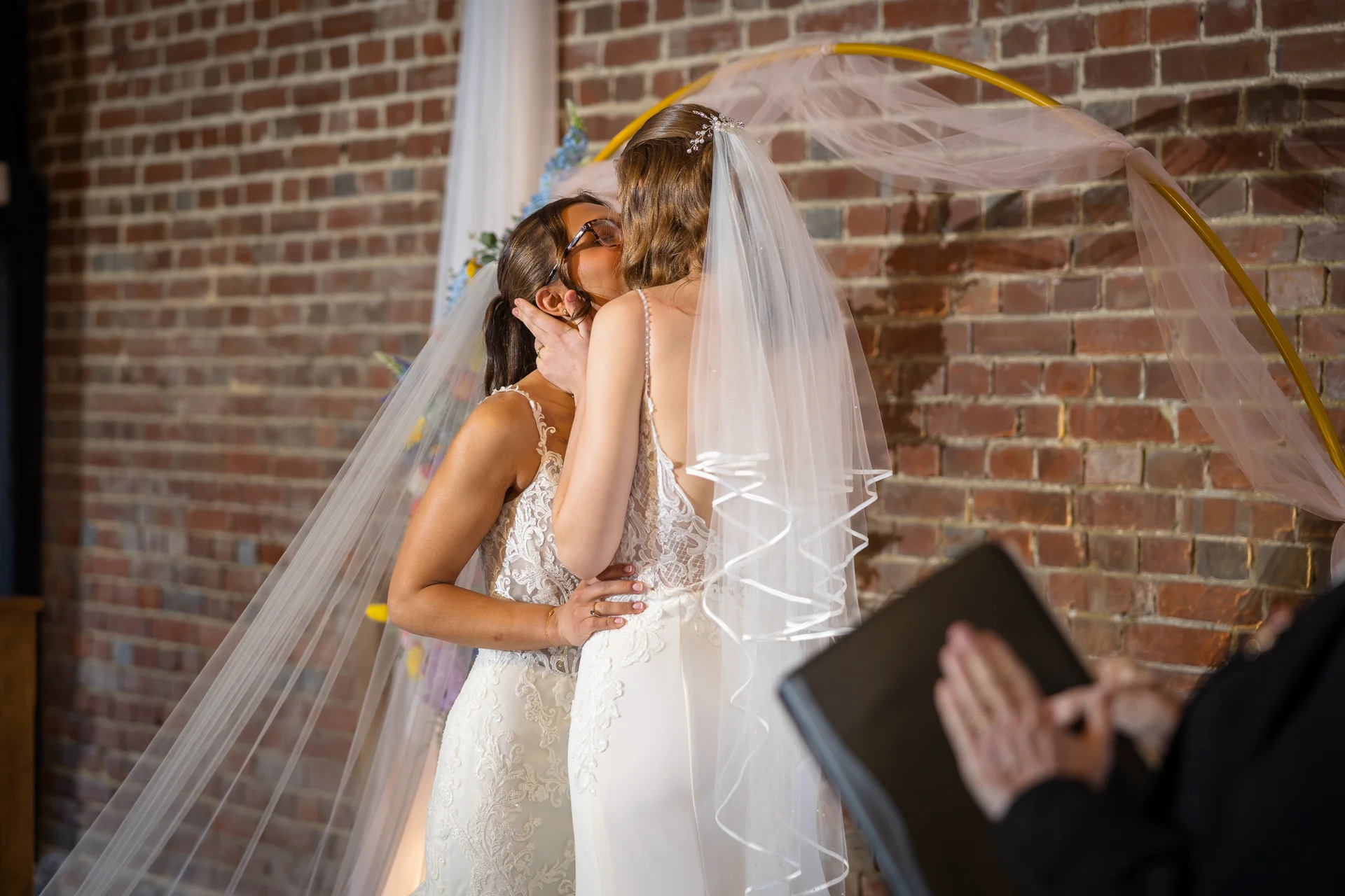Two brides share a tender first kiss under a golden ceremony arch against an exposed brick wall during their wedding ceremony.