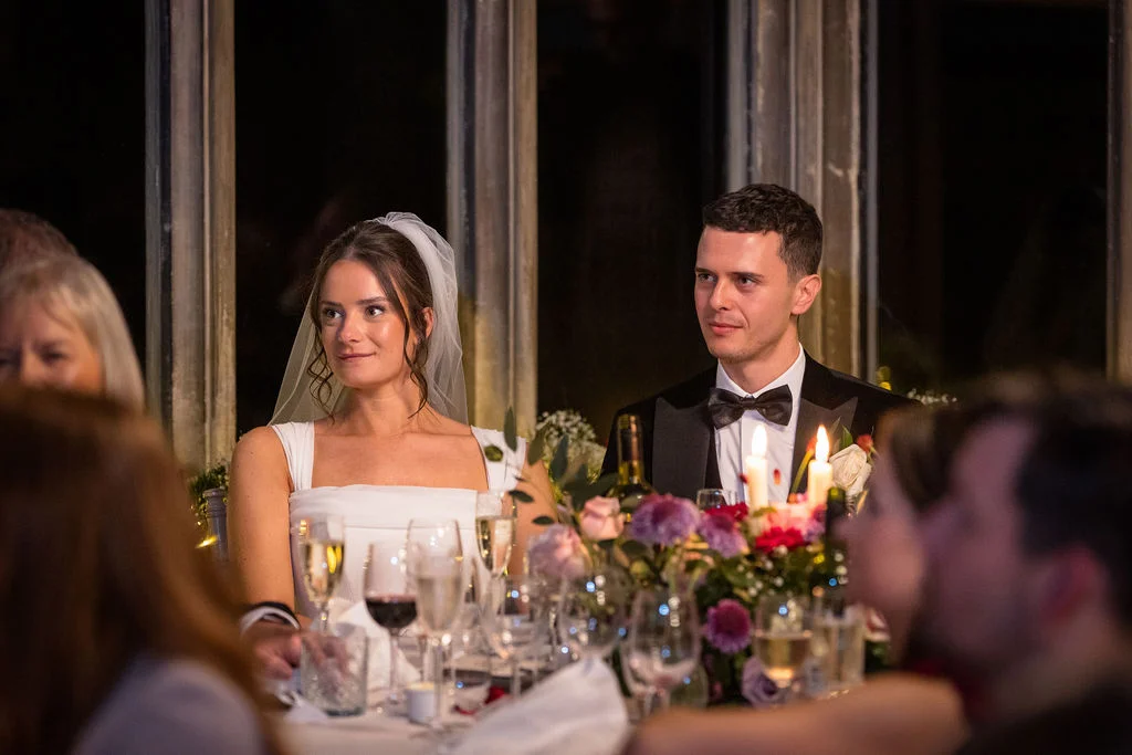 Newlywed bride and groom sitting at the top table during wedding speeches, surrounded by candlelight, floral arrangements, and guests.