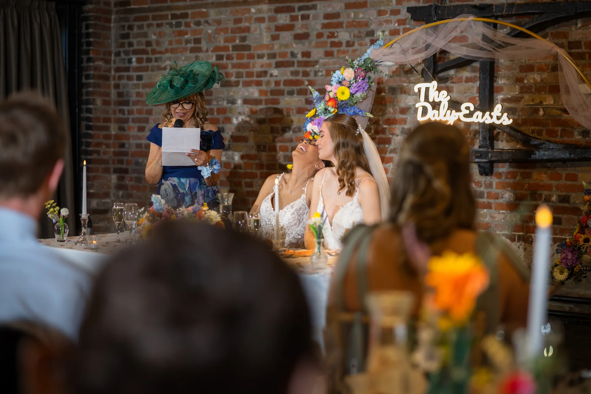 A mother of the bride in a dramatic green fascinator delivers a speech, making the two brides laugh joyfully at the top table in a rustic brick venue.