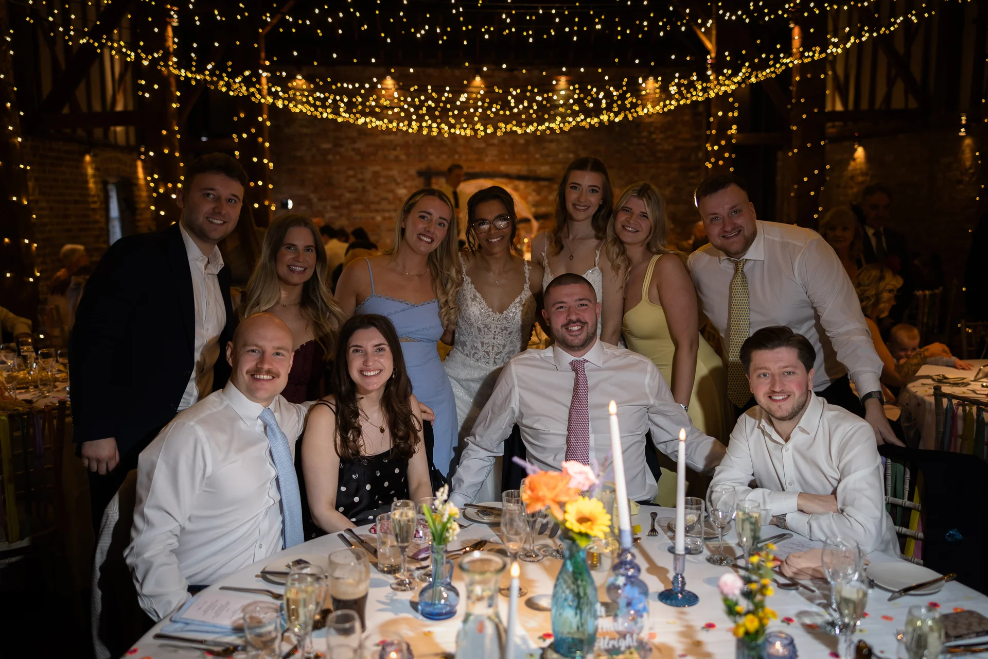 A large wedding party and guests gather around a candlelit dining table beneath a stunning ceiling canopy of warm fairy lights in a rustic brick barn venue.