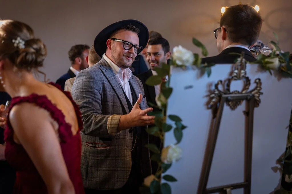A stylish guest wearing a checked blazer and hat chats animatedly with others near a decorated easel sign during a wedding reception.