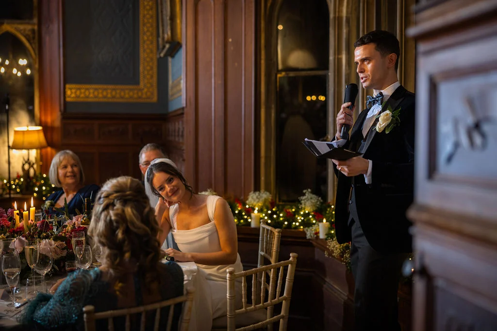 The groom delivers his speech wearing a black tuxedo with a white rose buttonhole while the smiling bride looks on affectionately from the candlelit head table.