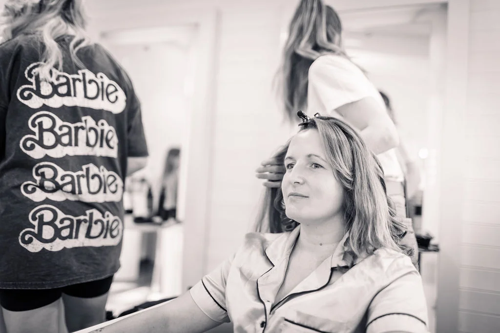 A black and white photograph captures a bride having her hair styled during morning preparations while wearing satin pyjamas, with a bridesmaid in a barbie-printed shirt standing nearby in front of mirrors.