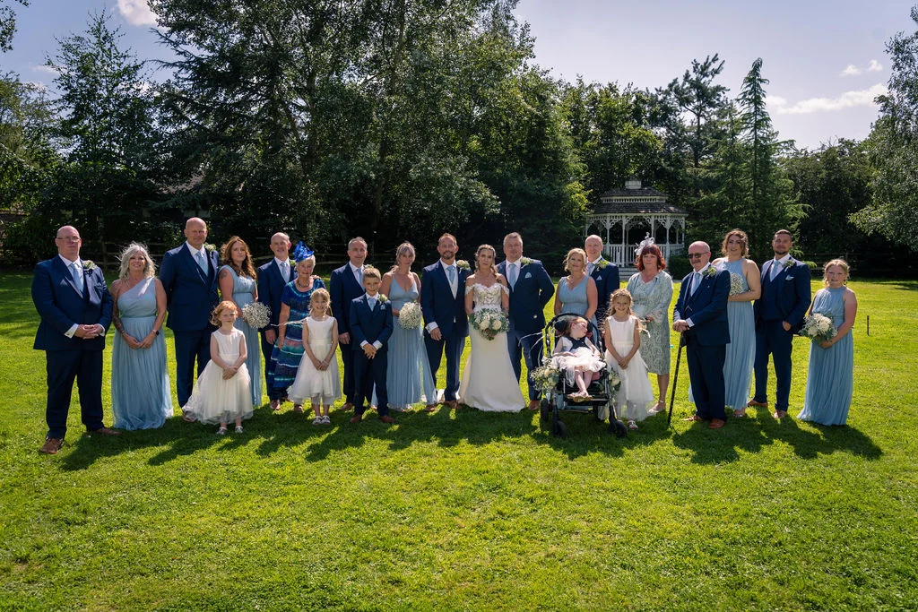 A large wedding party and family group pose on a sunlit lawn outdoors with a white gazebo in the background.