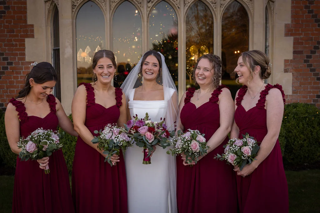 A joyful bride and her four bridesmaids in burgundy dresses stand laughing together in front of large arched windows holding floral bouquets.