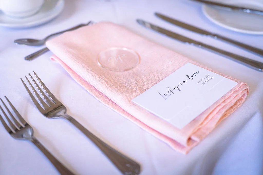 A close-up view of an elegant wedding table setting featuring a neatly folded pink napkin resting on white linen, accompanied by polished silver cutlery and a personalised stationery card.