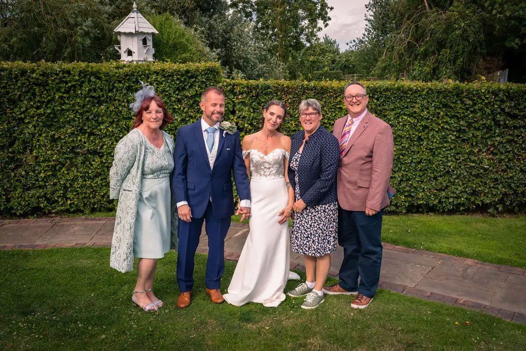 A newlywed couple poses outdoors in a manicured garden alongside family members against a tall green hedge.