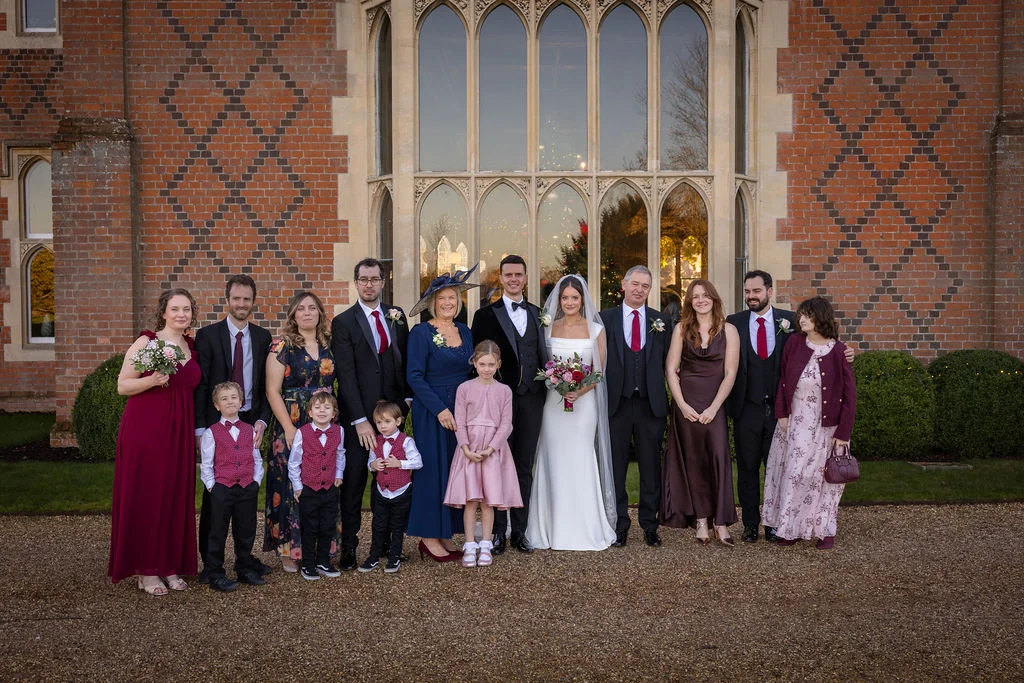 A large wedding party gathers outside a stately red brick country house with grand arched windows for a formal group portrait.
