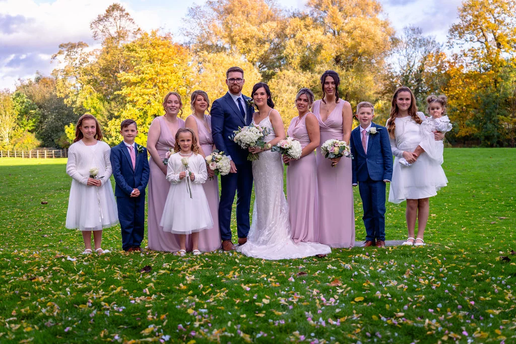 A newlywed couple poses outdoors with their large wedding party of bridesmaids in pink dresses, groomsmen, and children amidst a backdrop of vibrant autumn foliage and a green lawn scattered with fallen leaves.