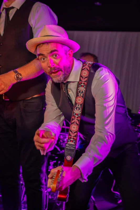 An energetic musician wearing a fedora and patterned guitar strap performs on stage under vibrant purple lighting during the evening wedding reception.