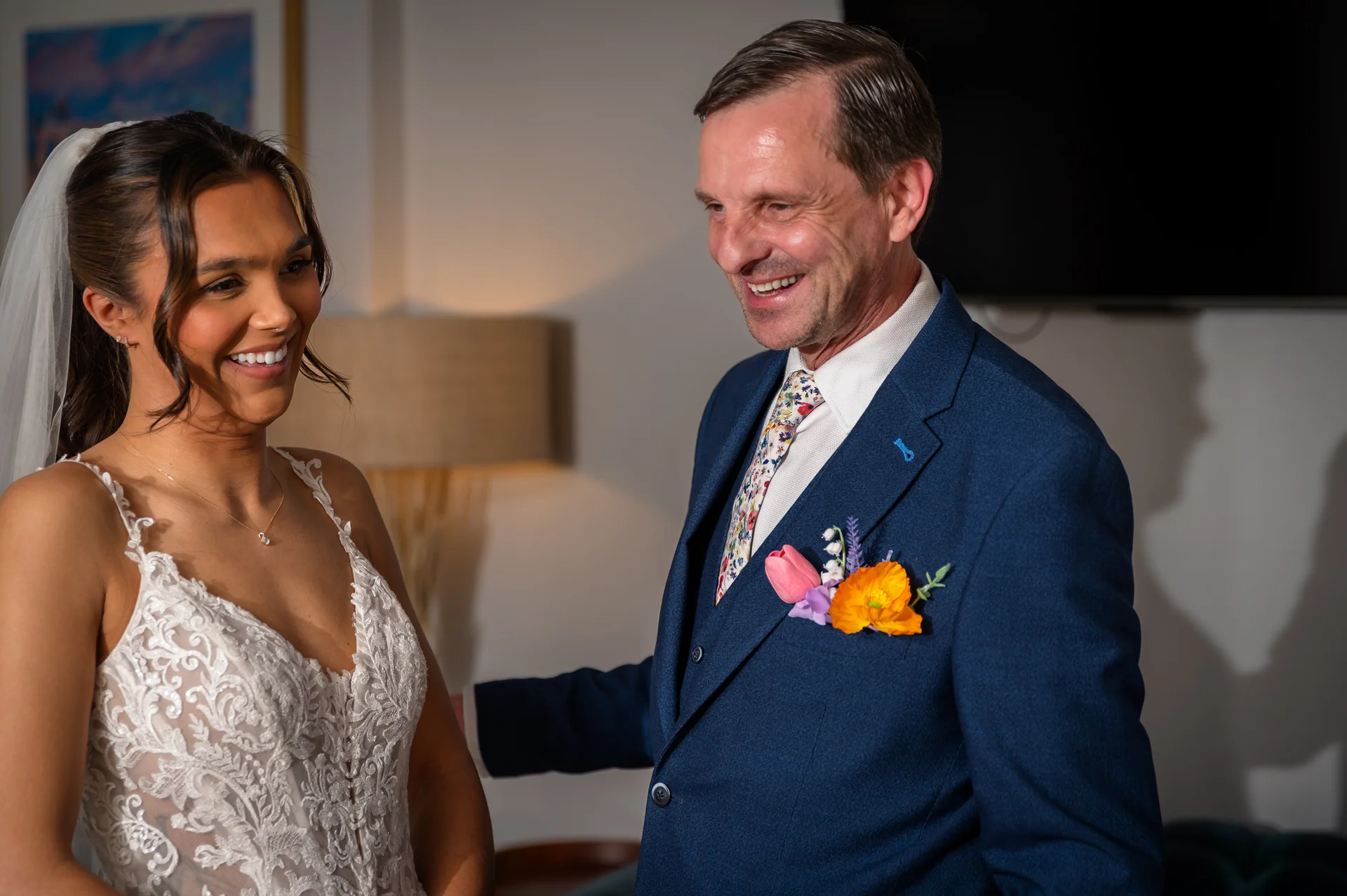 A smiling bride wearing a lace wedding dress shares a joyful moment with her father in a blue suit during morning preparations.