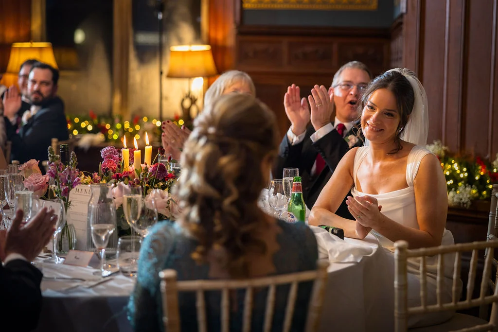 A smiling bride listens to wedding speeches at a candlelit top table while family members applaud in the background.