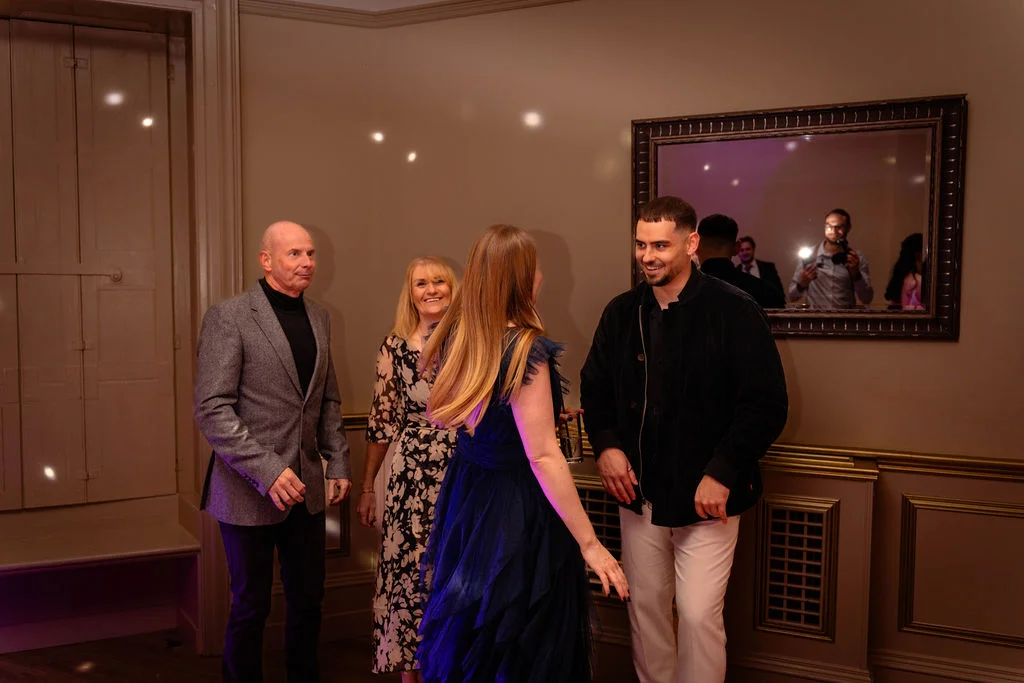 Wedding guests laugh and mingle together inside an elegant reception room, captured in a candid moment with a decorative wall mirror in the background.