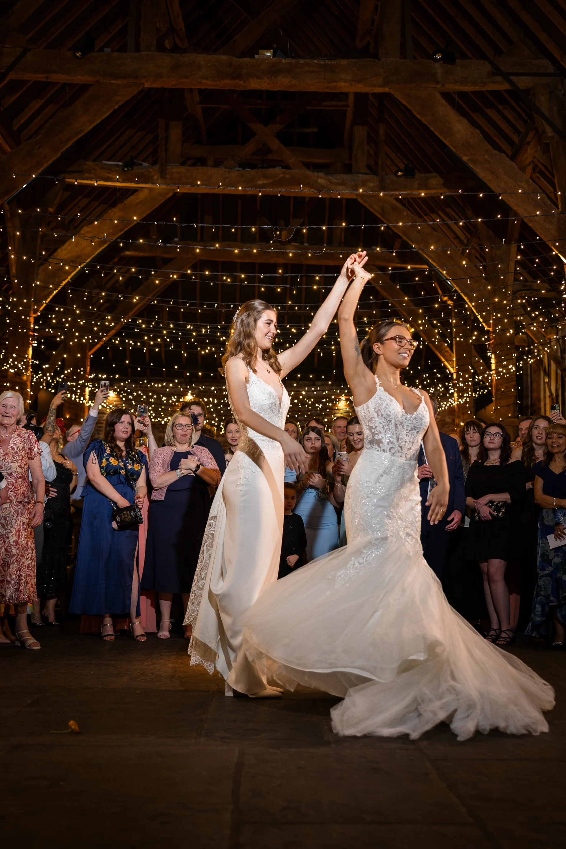 Two brides share their first dance inside a rustic barn illuminated by a canopy of string lights while guests watch and record.