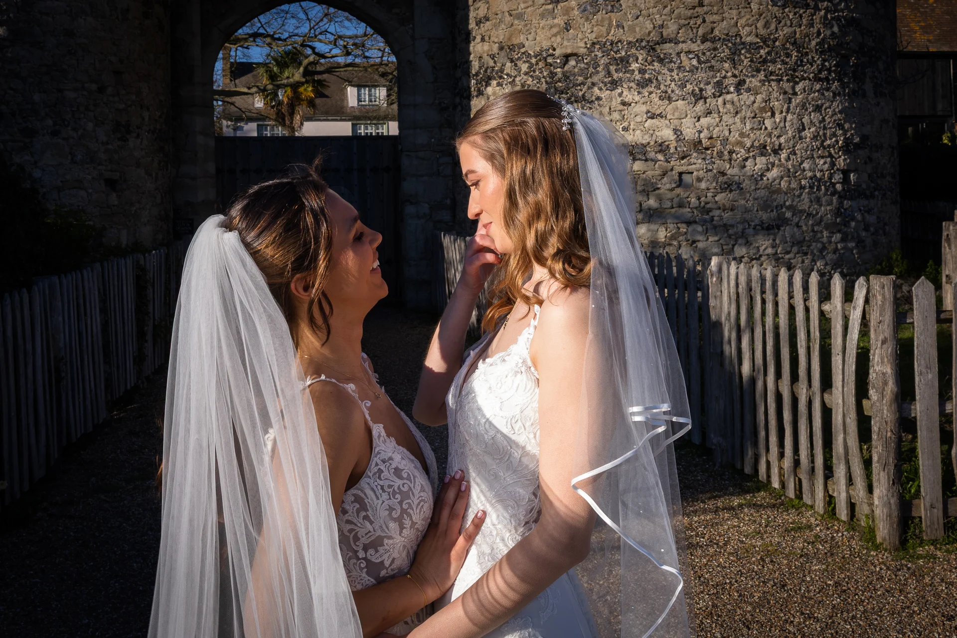 Two smiling brides in lace wedding dresses and veils share a tender moment standing outside against a historic stone archway during golden hour.