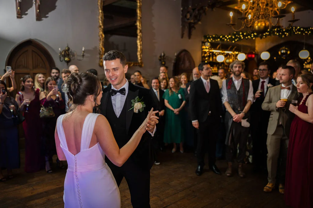 Newlyweds share their first dance in an elegant ballroom filled with watching guests under warm chandelier and fairy light illumination.