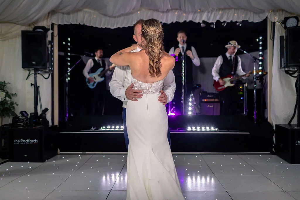 Newlyweds share their first dance on an illuminated white dance floor inside a marquee, with a live band performing on stage behind them.