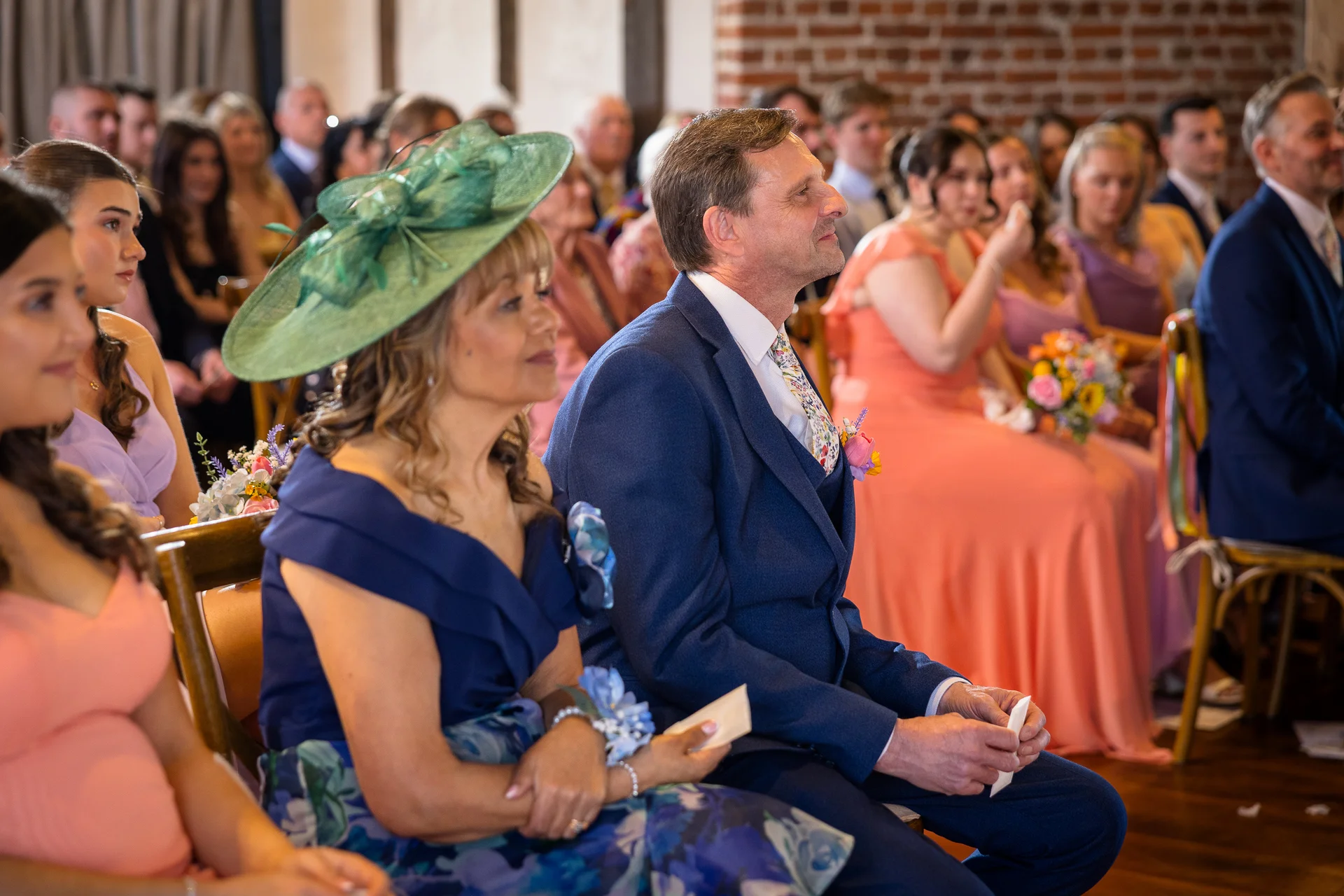 Guests seated attentively during a wedding ceremony in an indoor venue with exposed brick walls, featuring a woman in a blue outfit with a green fascinator and a man in a navy suit.