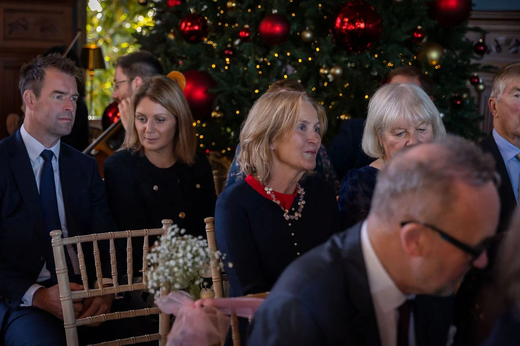 Wedding guests seated attentively during a ceremony with a large Christmas tree decorated with red baubles in the background.