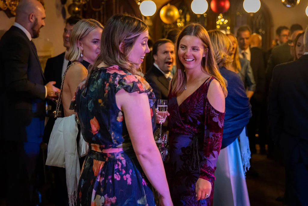 Wedding guests mingle and laugh together during a lively evening reception illuminated by warm overhead lights.
