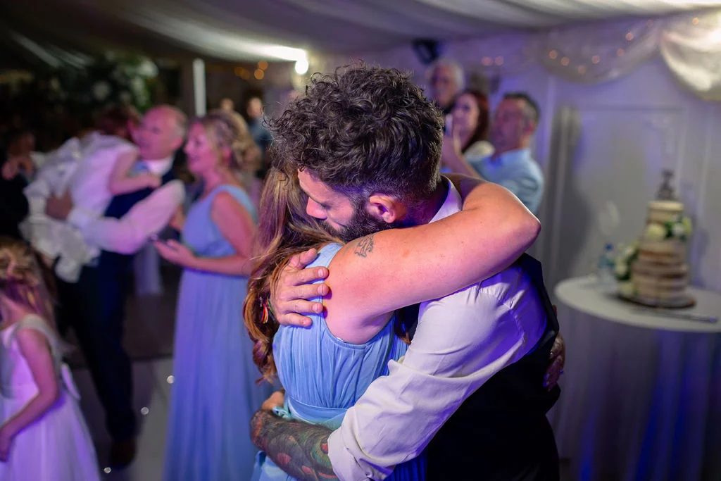 A bride and groom share a tender, emotional embrace in a softly lit marquee while guests mingle in the background during the evening reception.