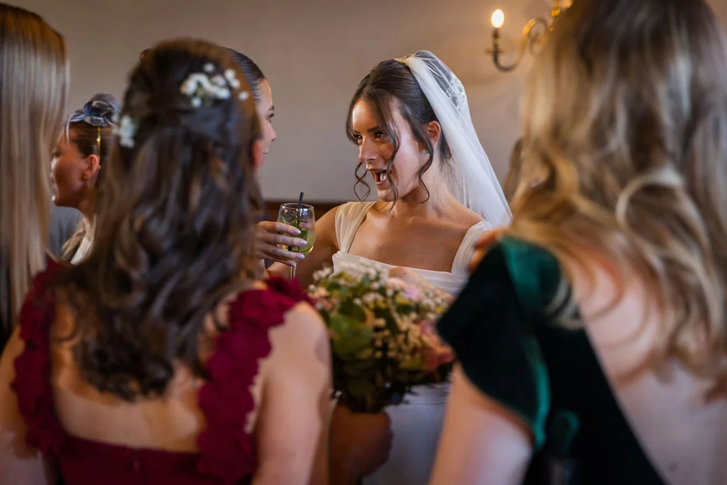 A joyful bride laughs and holds a cocktail while chatting with her bridesmaids during the drinks reception.