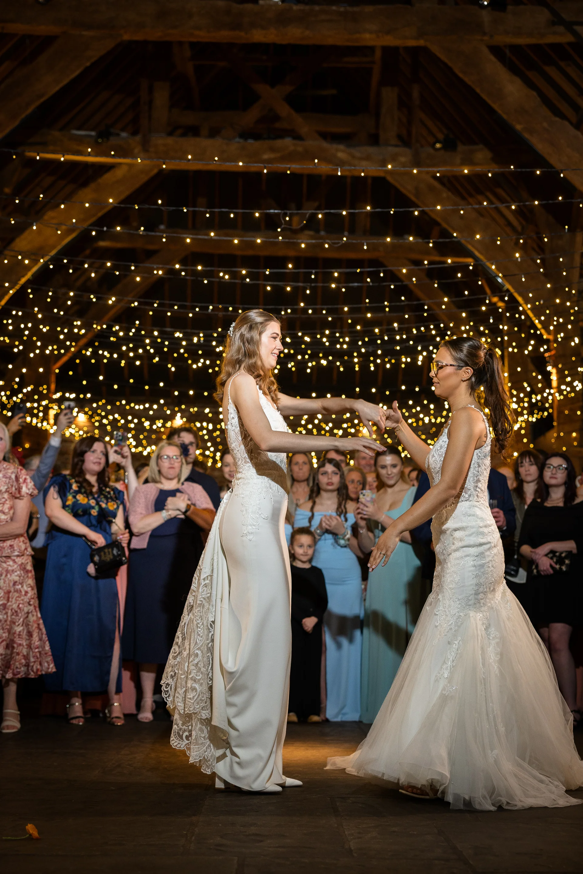 Two brides share their first dance inside a rustic barn illuminated by a dramatic canopy of fairy lights while guests watch on joyfully.