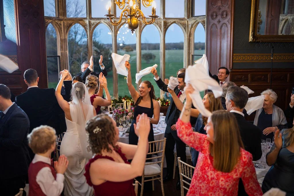 Wedding guests enthusiastically wave their napkins in the air during speeches inside a grand, wood-panelled stately home dining room with large arched windows.