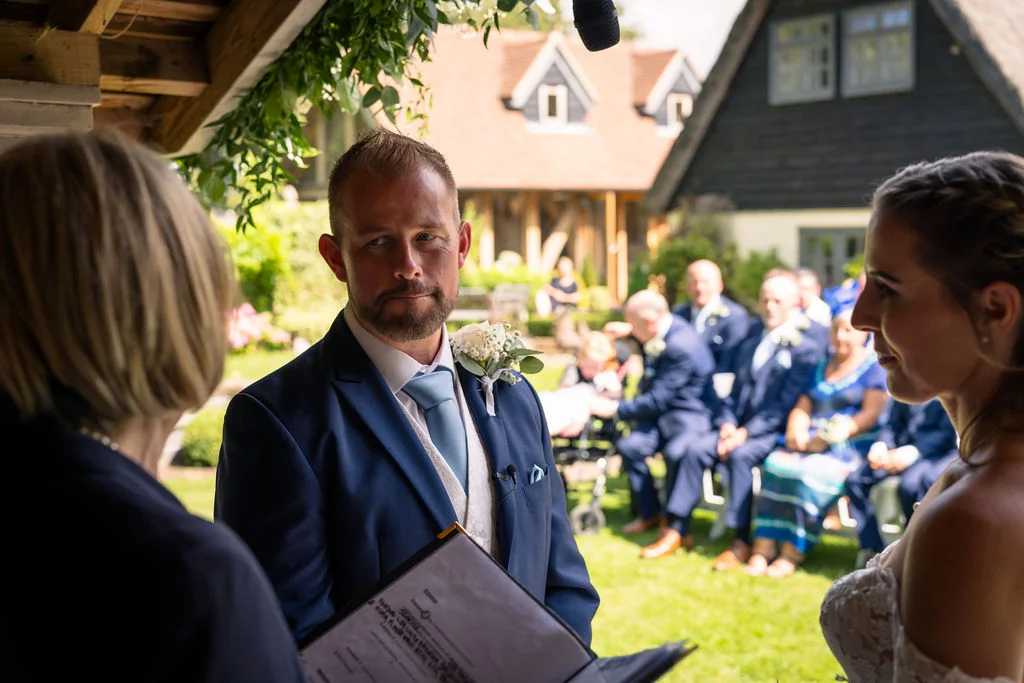 A groom in a navy suit and light blue tie stands attentively during an outdoor celebrant-led wedding ceremony, with guests seated on the lawn in the background.